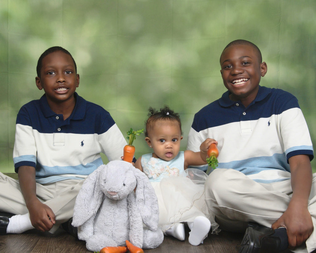 Two young black boys wearing Tommy Hilfiger striped polos smile while sitting on either side of their infant cousin who is wearing a blue and white dress and holding carrots in both her hands