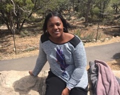 A black woman wearing a gray long sleeve shirt sits on a rock wall next to jackets
